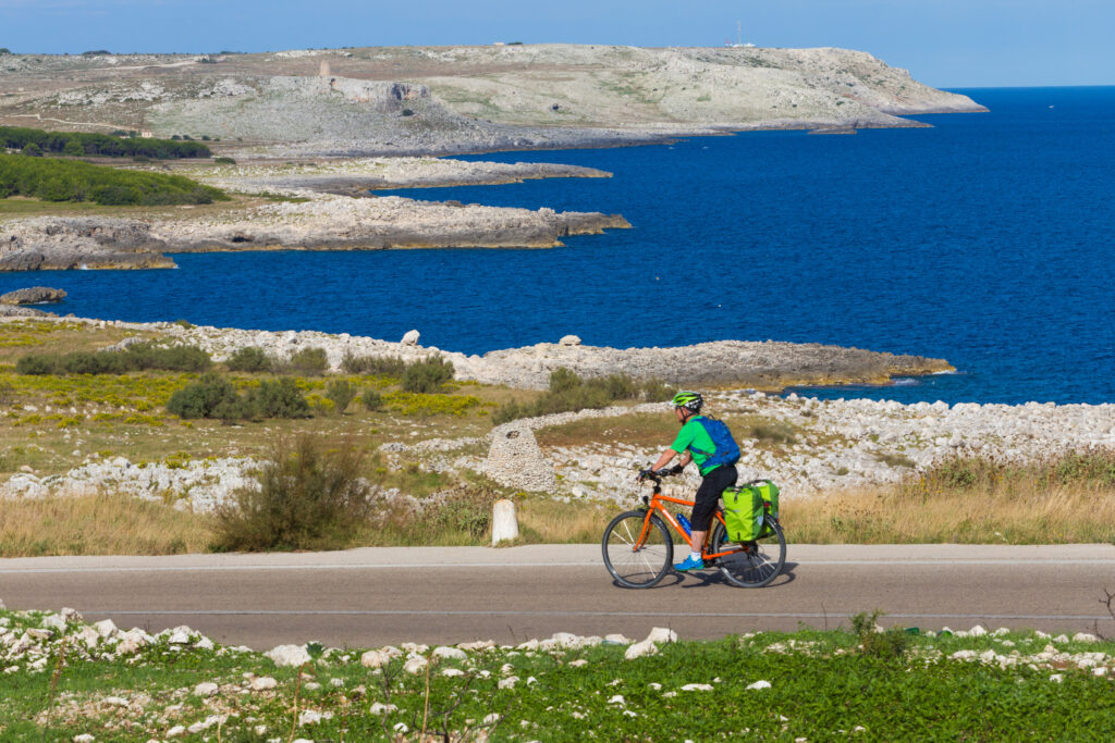 Ciclista in viaggio con "Girolibero" pedala lungo un sentiero costiero in Salento con panorama sul mare